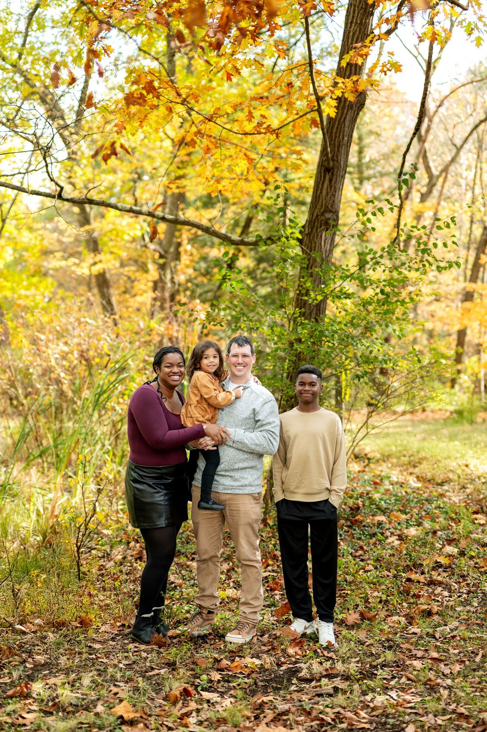 A joyful interracial family of four poses among colorful autumn foliage in a wooded park setting.