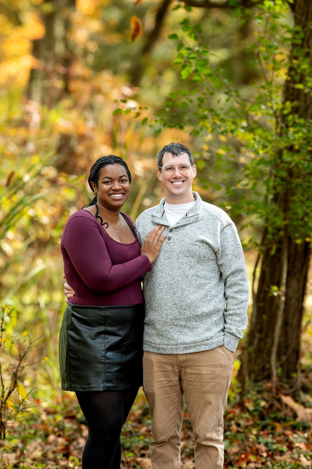 A smiling interracial couple stands together in a forest with vibrant fall foliage in the background.