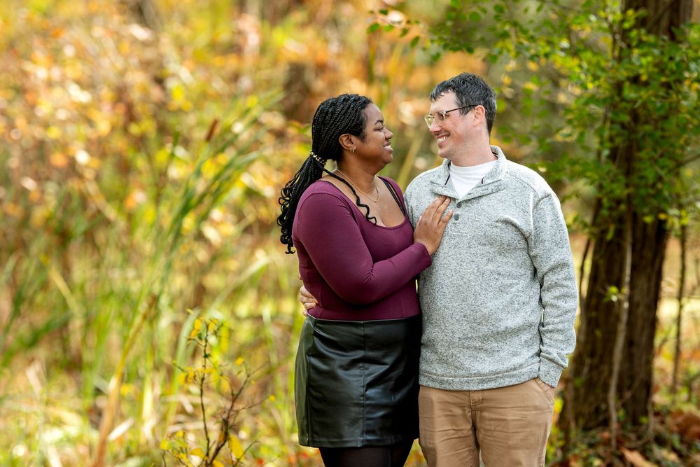 An interracial couple gazes lovingly at each other surrounded by golden autumn leaves in a forest.