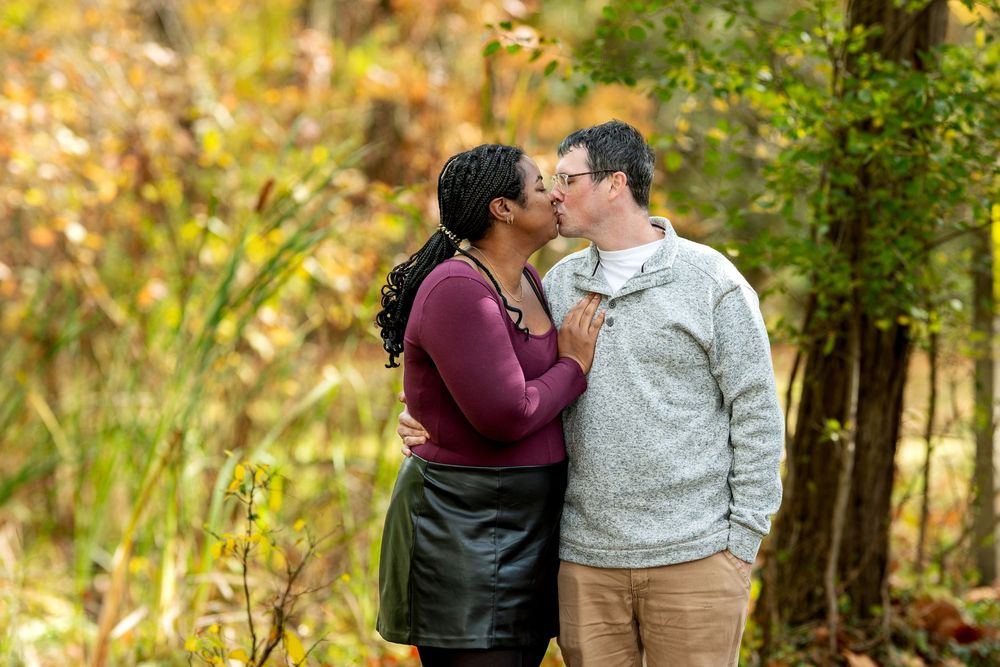 An interracial couple shares a tender kiss outdoors among warm fall foliage in a wooded setting.