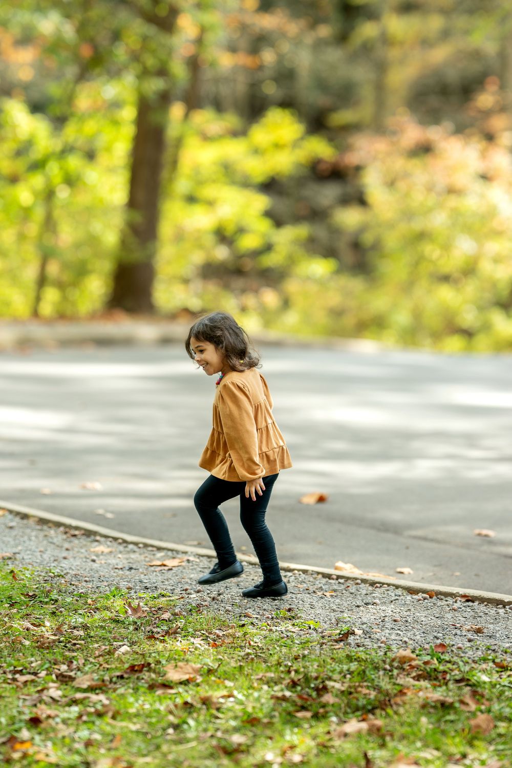 A young girl in an orange dress playfully poses on a path surrounded by lush autumn greenery.