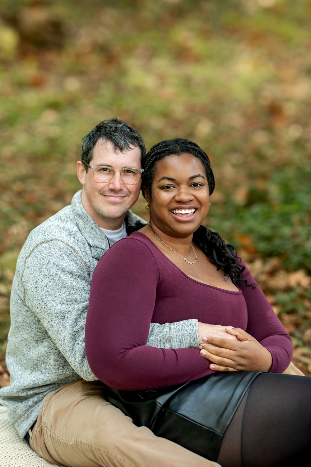 A happy interracial couple sits together on the ground smiling in a colorful autumn forest setting.