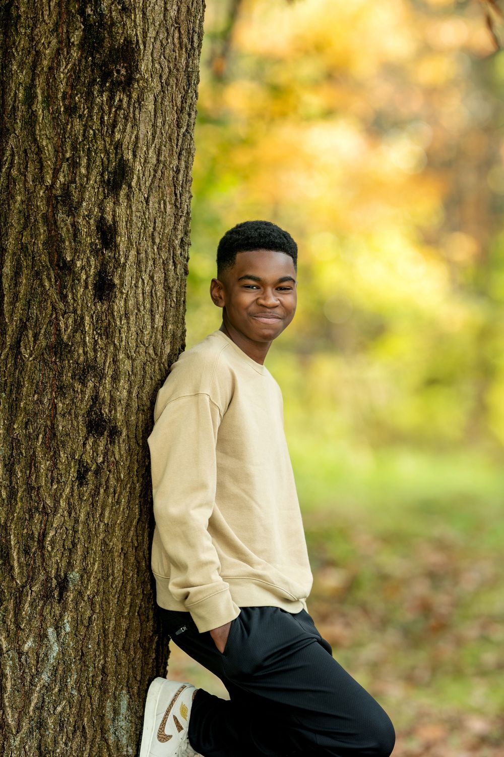 Smiling young Black teen leaning against a tree trunk in a beige sweatshirt during autumn.