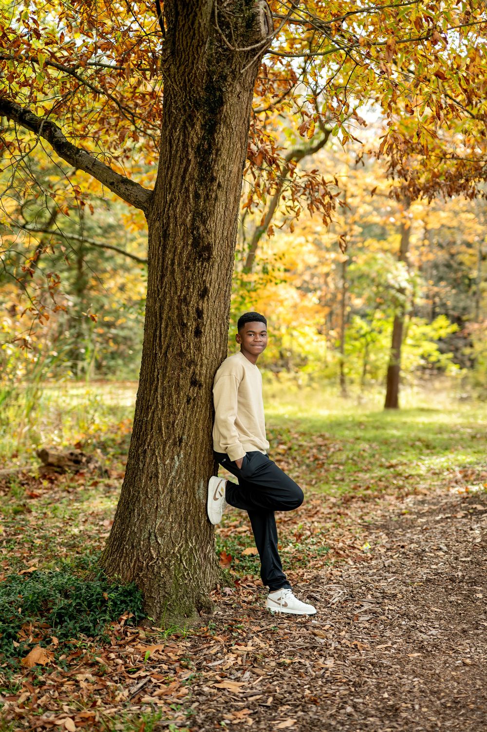 Young Black teen in beige sweater leaning against a tree on a fall foliage forest path.