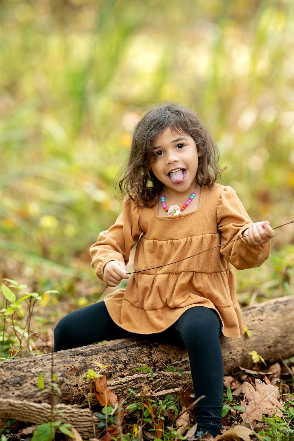 Playful little girl in orange dress sticking tongue out while sitting on a log in autumn woods.