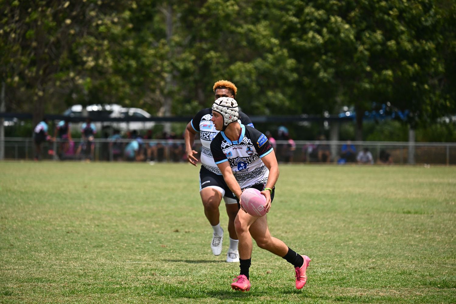 Rugby player in pink cleats runs with the ball during a match on a grassy field with spectators in background.