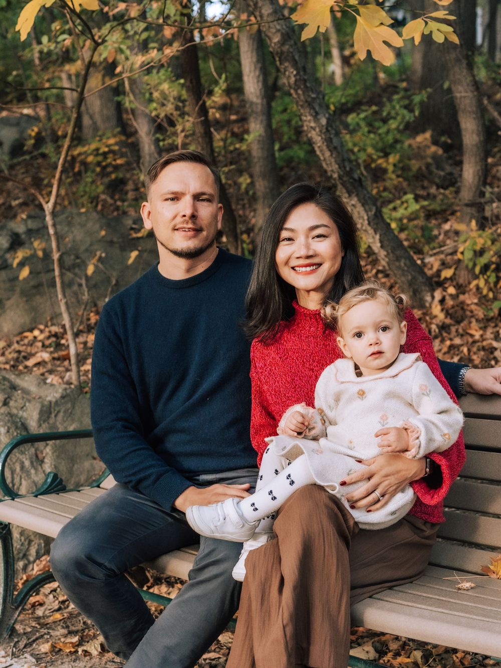 A family sits together on a wooden bench surrounded by autumn foliage at Menotomy Rocks Park in Arlington, MA.