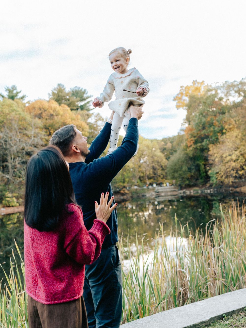 A couple lifts and plays with their baby outdoors against a backdrop of cattails and autumn trees.