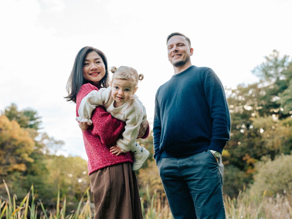 A family shares joyful moments together outdoors during a fall photo session near a pond, Menotomy-rocks-park in Arlington, MA.