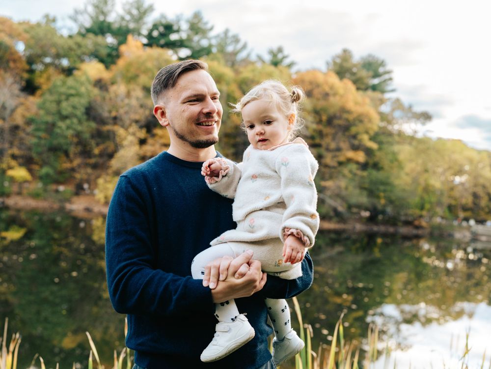 Family embraces while standing near a serene lake surrounded by autumn trees and golden sunlight, Menotomy-rocks-park in Arlington, MA.
