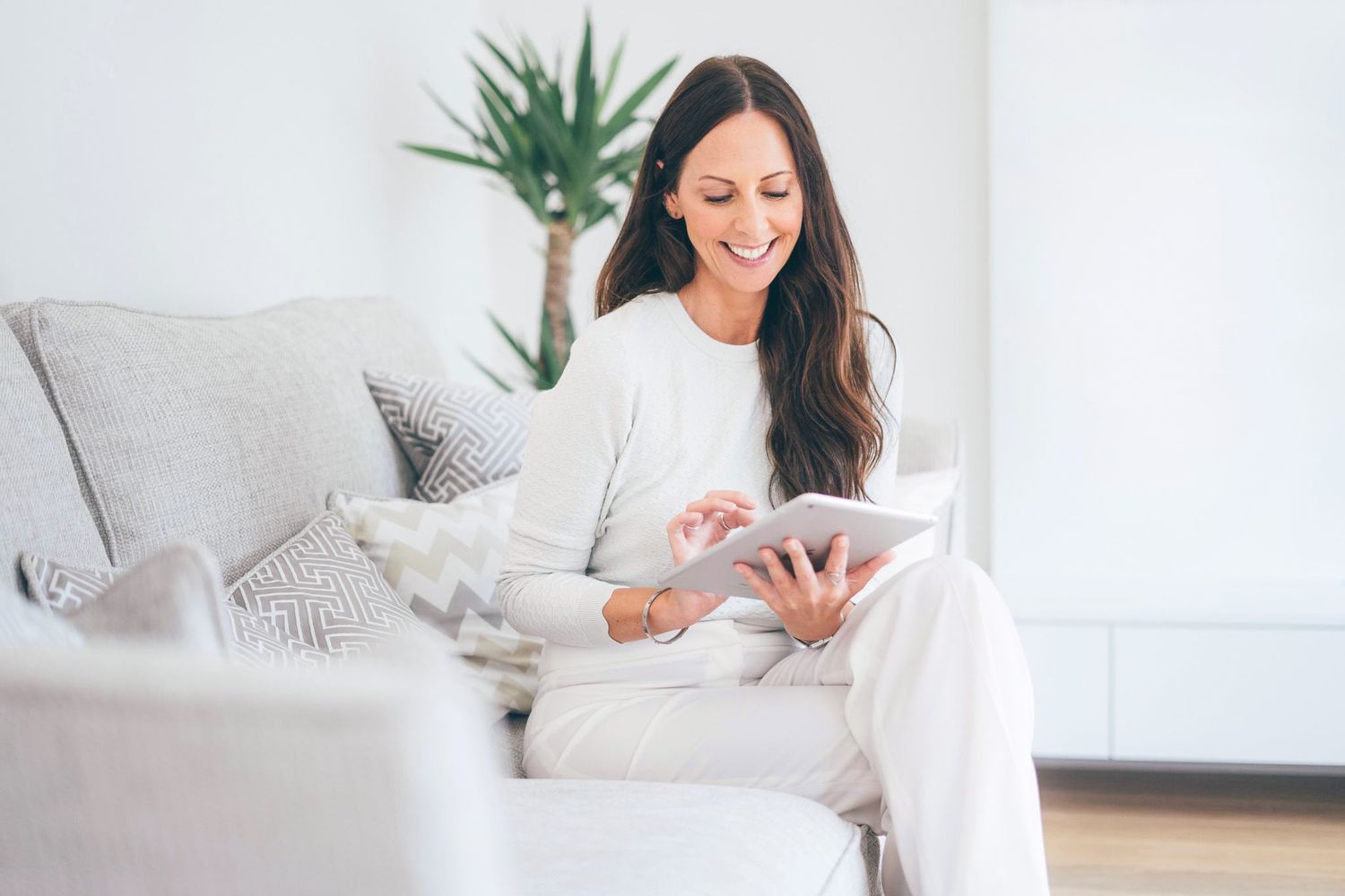 A person in white loungewear relaxes on a light gray couch while using a digital tablet in a bright modern room.