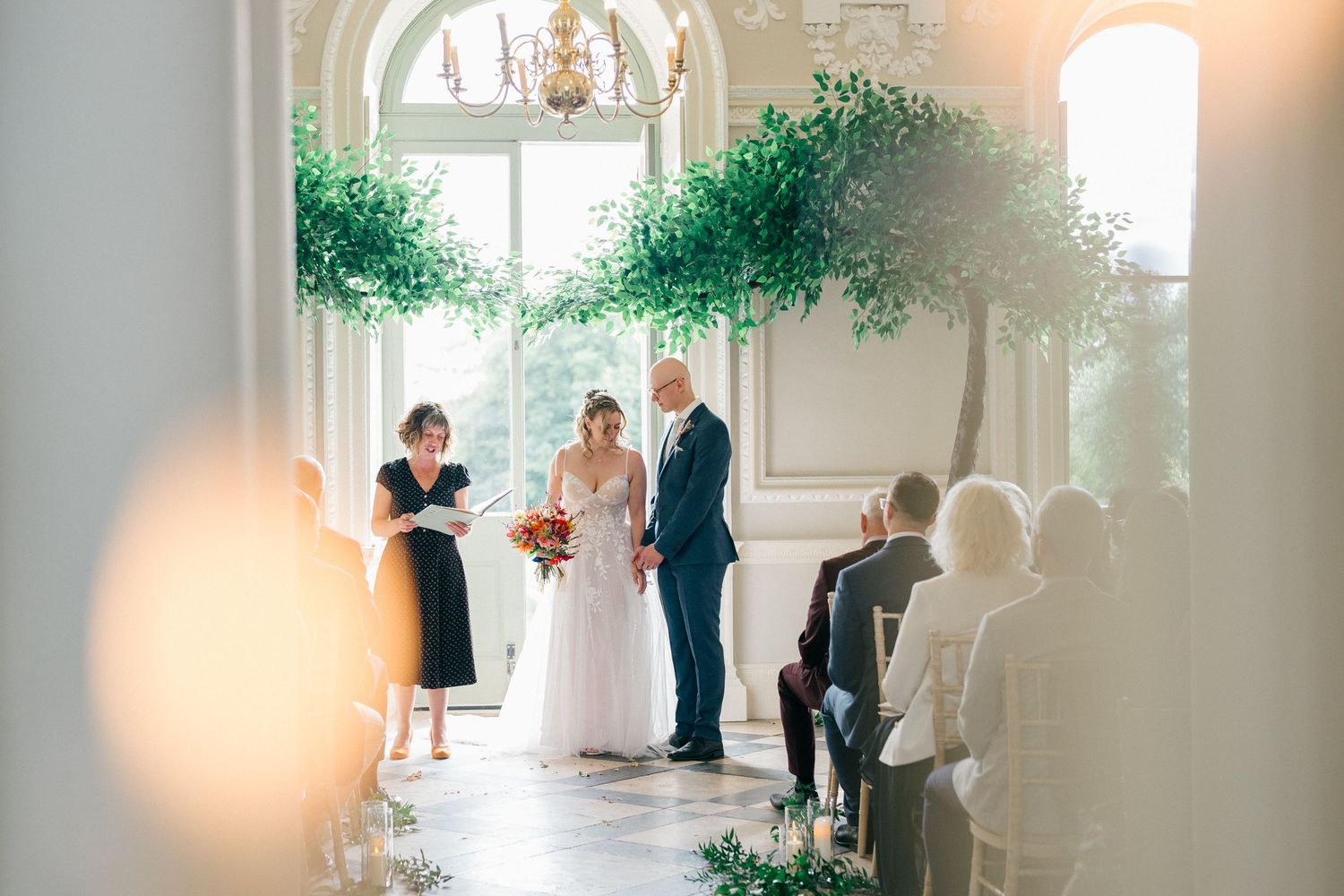 Elegant wedding ceremony scene in a bright white room with large windows and green foliage decorations for Nova Scotia wedding photographer.