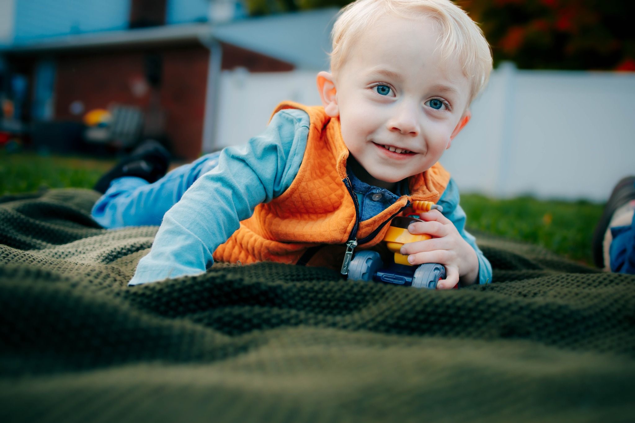 Baby wearing orange vest sitting on dark blanket outdoors.