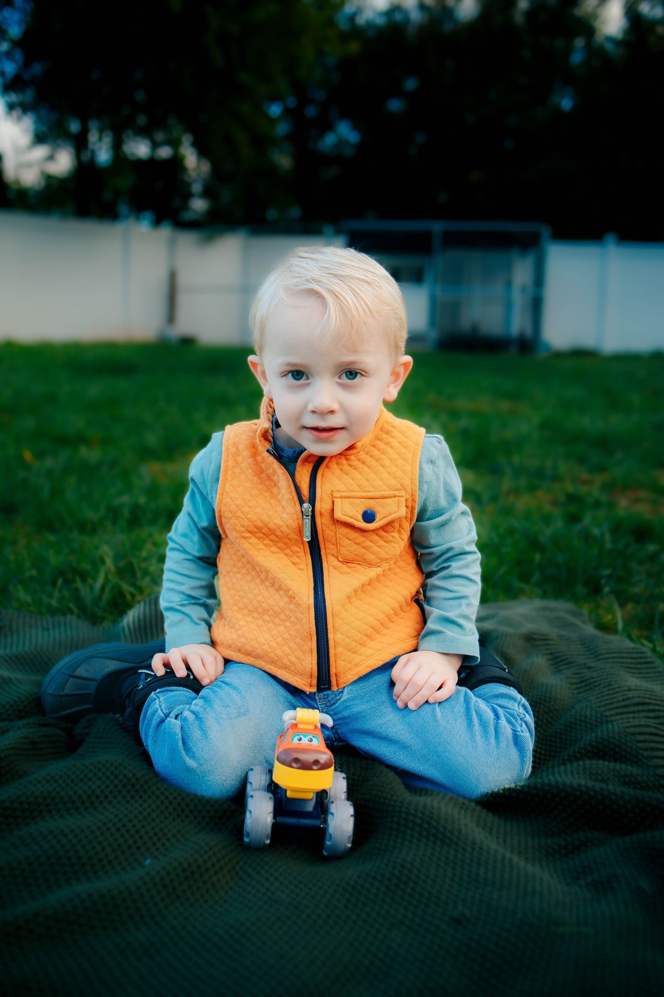 Baby in orange quilted vest and gray shirt playing on blanket in grass.