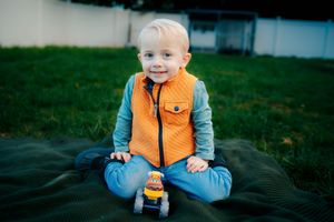 Baby wearing orange puffy vest smiling while sitting on blanket outside.