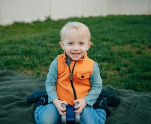 Young child sitting on a blanket outdoors wearing an orange vest and jeans.