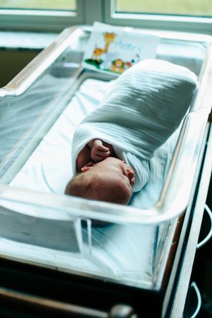 Medical staff examines newborn baby in hospital bassinet through protective plastic covering.