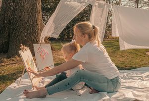 Two people sit together on a white blanket outdoors while drawing or writing in notebooks on a sunny day.