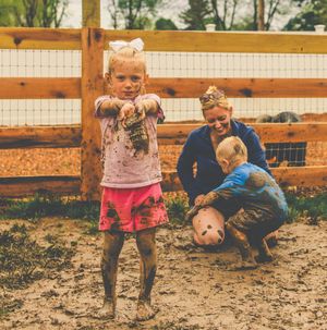 Children playing together in muddy ground near a wooden fence on a sunny day.