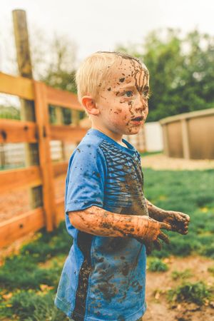A young child in a blue shirt covered in mud plays outdoors near a wooden fence.