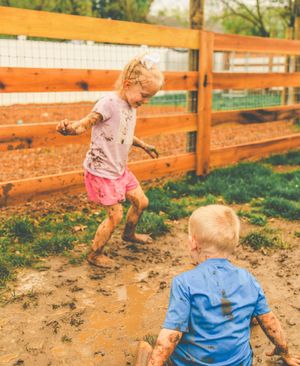 Children playing and splashing in muddy puddles on a sunny day near a wooden fence.