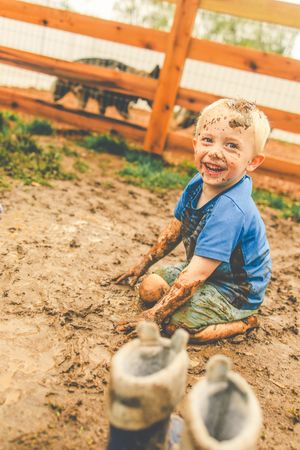 A muddy outdoor scene shows a child in blue playing in the dirt with boots nearby on a sunny day.