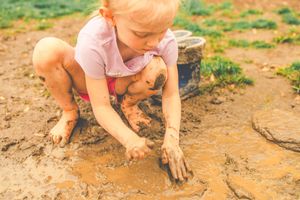 Small child in pink shirt plays in muddy dirt on a sunny day while exploring nature outdoors.