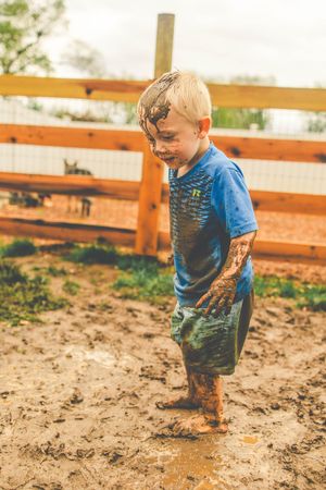 Young child playing and getting muddy while wearing blue shirt in a fenced dirt area on a sunny day.