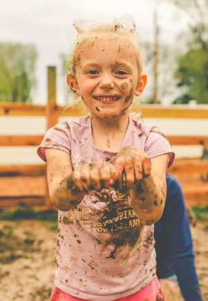A child in a pink shirt plays joyfully in the mud, showing their muddy hands and smiling face while playing outdoors.