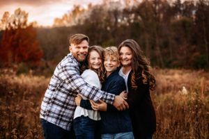 A family of four embraces while standing in a golden autumn field at sunset.