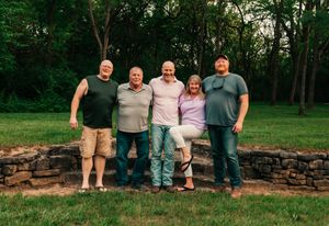 A group of people sitting casually on a rustic stone wall in an outdoor garden setting.
