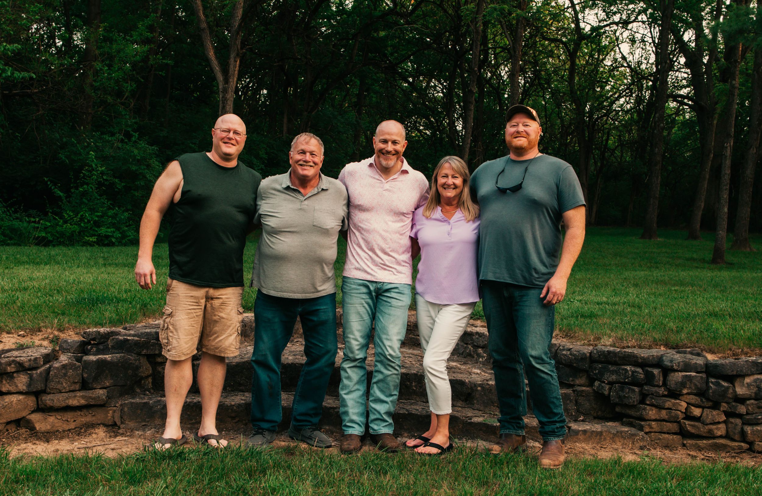 A group of people sitting together on a rustic stone wall surrounded by lush green trees and grass.