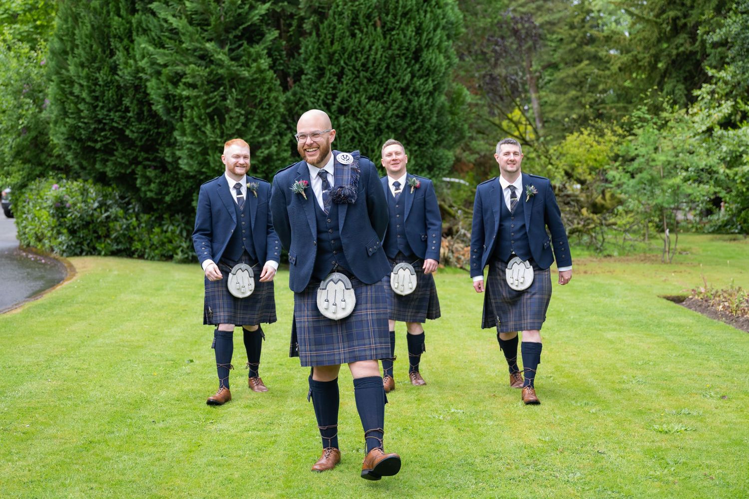 Groomsmen in navy suits walk together on a lush green lawn during an outdoor wedding celebration.