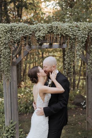 Bride and groom share first kiss during wedding ceremony at Farmer Brown's Greenhouse as the sun sets behind them, outdoor fall wedding