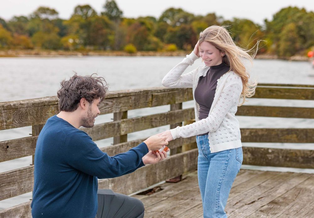 Man kneeling on wooden pier proposing to smiling woman along the great bay shoreline in Durham NH with 
autumn trees in background.