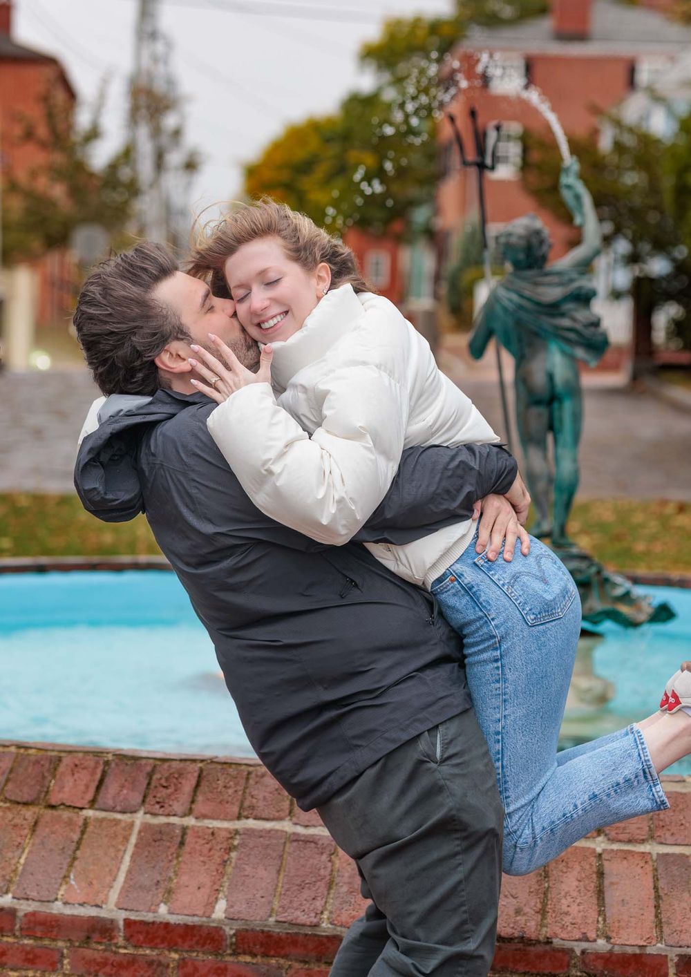 A couple shares a romantic kiss near a fountain with a bronze statue, as the man lifts the woman in a joyful embrace.
