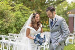 Smiling bride and groom in gray suit share joyful moment holding blue bouquet at outdoor ceremony.