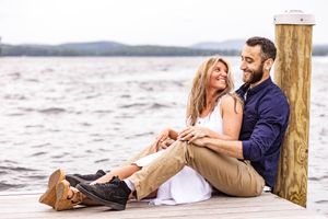 Couple sitting on a lakeside dock In Alton NH, smiling and embracing with mountains in the background.