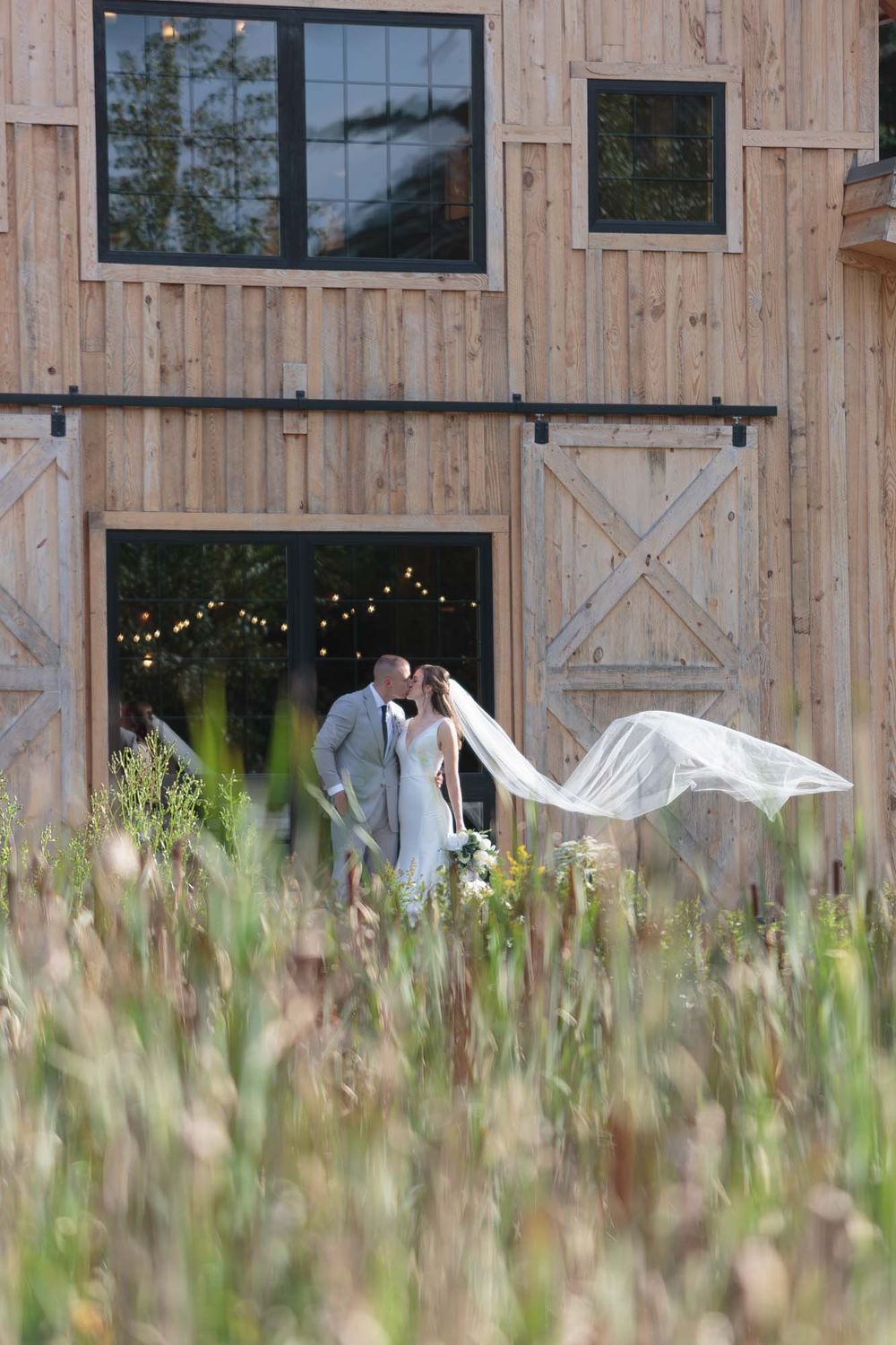 Bride and groom kissing in front of a wooden barn with veil flowing in the wind.