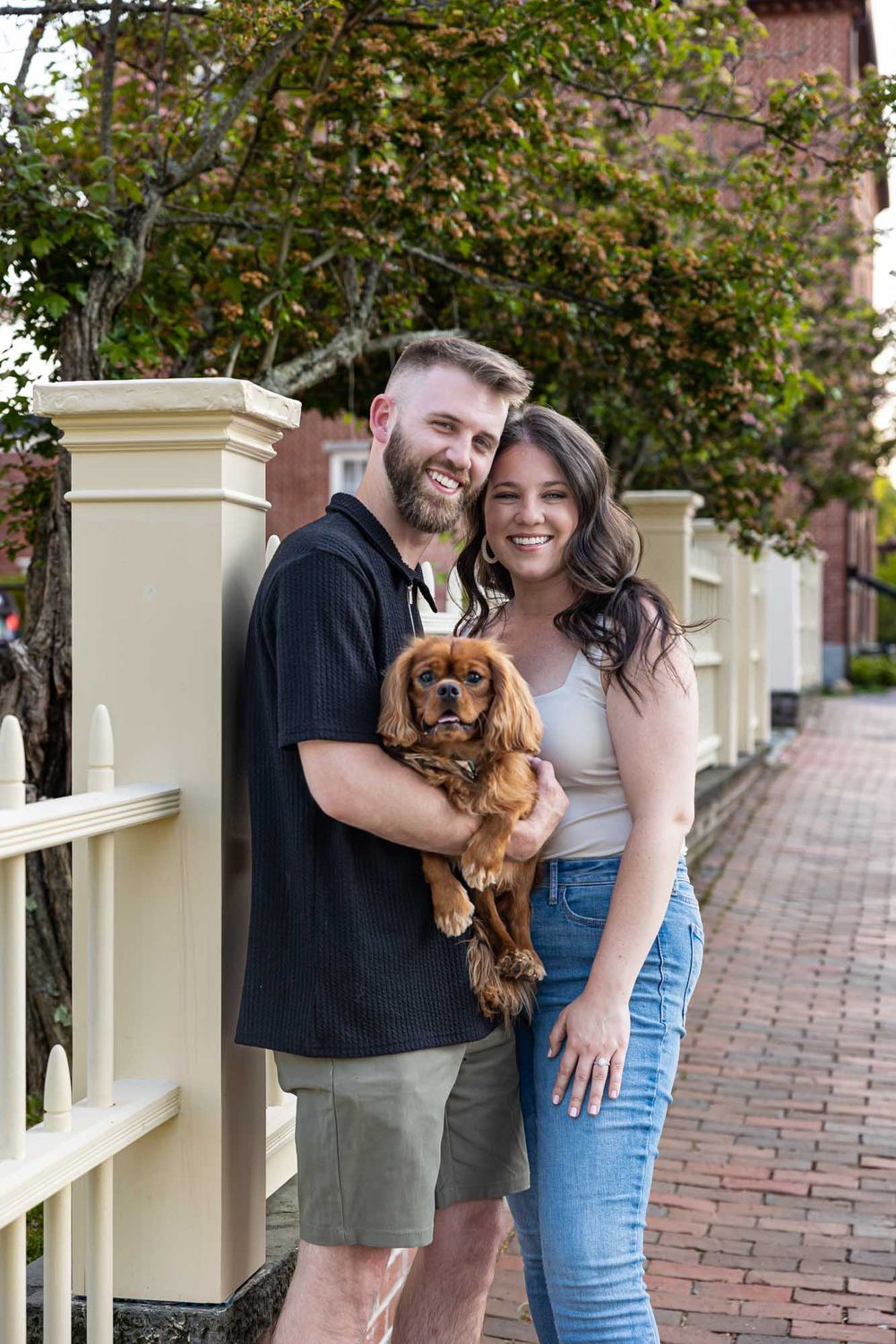 A smiling couple poses with their adorable Cavalier King Charles Spaniel on a charming brick sidewalk.