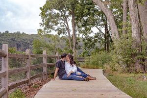 A romantic couple sits close together sharing a kiss on a wooden boardwalk surrounded by lush trees. Wagon Hill Farm Durham NH.