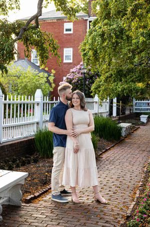A couple embraces on a brick path beside a white picket fence and red brick building surrounded by lush greenery.