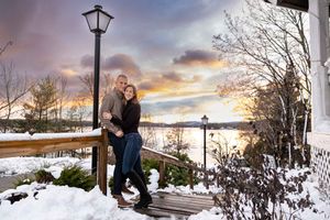 A happy couple embraces on snowy steps at sunset with a beautiful winter lake Winnipesaukee view behind them in Wolfeboro NH.