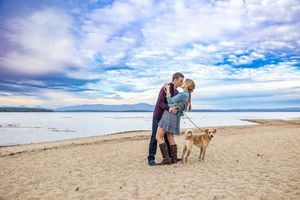Engaged Couple kissing on a sandy beach In Wells Maine with their golden dog under a dramatic York county cloudy sky. in Southern ME.