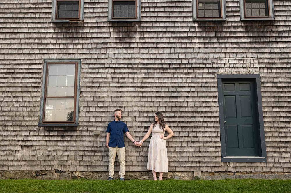 Couple holding hands in front of a rustic shingle building with blue door during an outdoor engagement session.