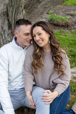 A happy couple sits together against a tree trunk in a park, sharing a warm and joyful moment.