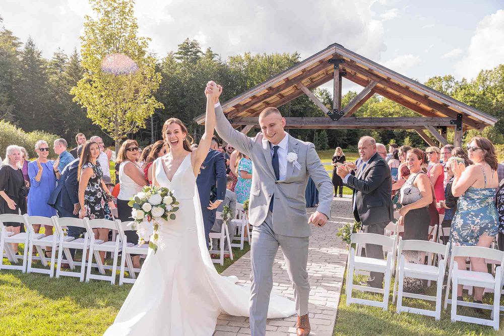 Bride and groom celebrate recessional at outdoor wedding ceremony with guests cheering with white chairs and a wooden pavilion in Concord NH