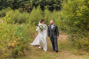 A bride and groom walk hand-in-hand along a wooded path, bride holding a white bouquet, both dressed in wedding attire.