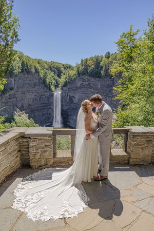 Bride and groom kiss in front of stunning Taughannock Falls waterfall under clear blue summer sky.