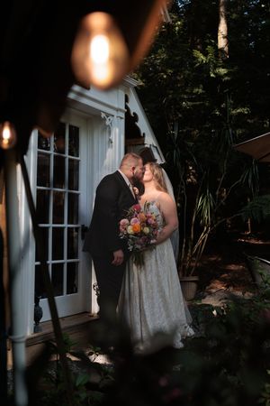 Bride and groom share romantic kiss outside charming white cottage surrounded by lush garden at dusk. Tarbin Gardens Franklin NH